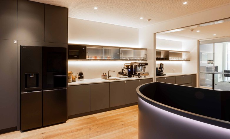 Moderne Büroküche mit dunklen Einbauschränken, integrierter Kühl-Gefrierkombination und heller Arbeitsfläche. Mehrere Kaffeemaschinen, Spüle und offene Regale mit Gläsern stehen für die Nutzung bereit. Im Vordergrund befindet sich ein geschwungener Tresen. Warme Beleuchtung, Holzboden und ein offener Durchgang zu weiteren Büroräumen schaffen eine hochwertige, einladende Atmosphäre.
