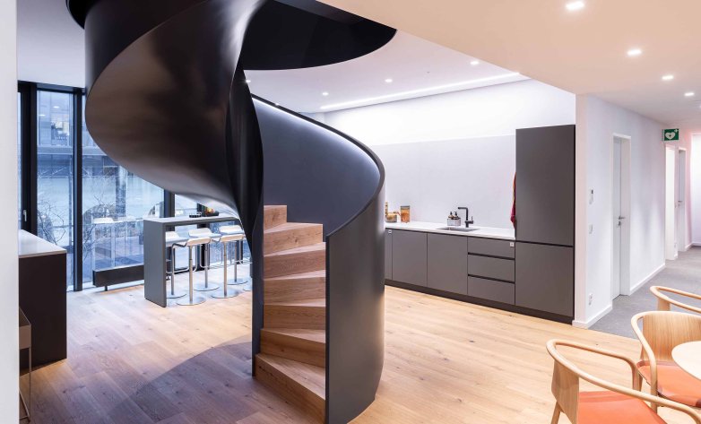 Offener Büro- und Aufenthaltsbereich mit einer markanten schwarzen Wendeltreppe als zentrales Gestaltungselement. Eine moderne Küchenzeile mit Spüle und Schränken befindet sich im Hintergrund, links ein Hochtisch mit Barhockern an der Fensterfront. Heller Holzboden, dezente Beleuchtung und eine offene Raumgestaltung schaffen eine moderne, einladende Arbeitsatmosphäre.