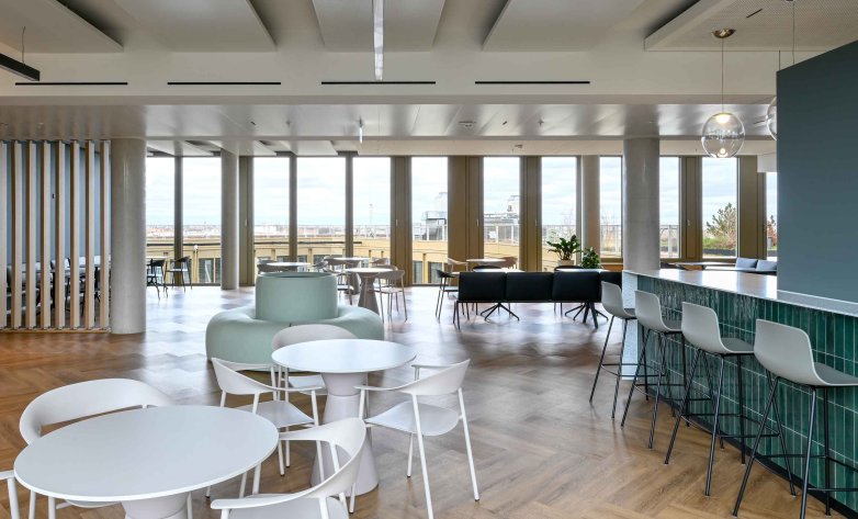Offener Küchen- und Loungebereich mit stilvoller Bar, großzügigen Fensterfronten und viel Tageslicht. Hochwertige Materialien, moderne Pendelleuchten und komfortable Sitzgelegenheiten schaffen einen einladenden Treffpunkt für Pausen, informelle Gespräche und den gemeinsamen Austausch im Arbeitsalltag.
