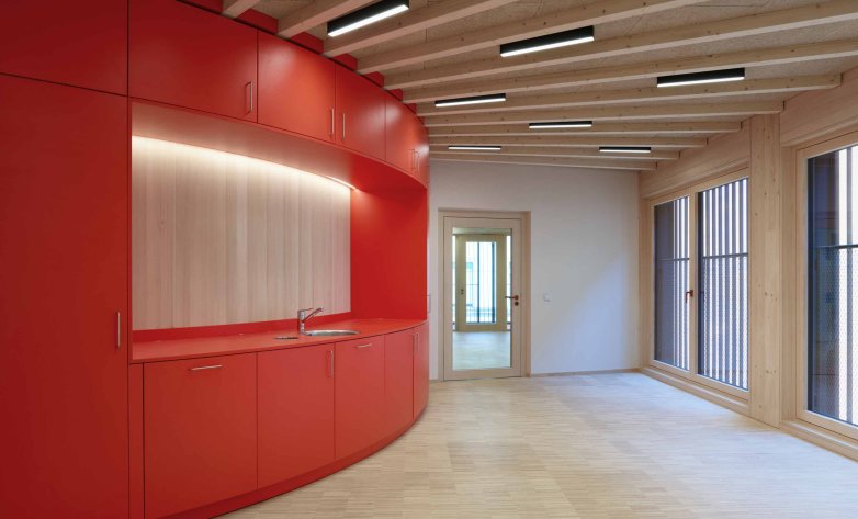 Heller, moderner Innenraum mit einer geschwungenen Küchenzeile in kräftigem Rot. Die Einbauten kombinieren rote Fronten mit einer hellen Holzrückwand und indirekter Beleuchtung. Sichtbare Holzbalken an der Decke sowie der helle Holzboden schaffen eine warme Atmosphäre. Große Fenster mit vertikalen Lamellen lassen viel Tageslicht herein.