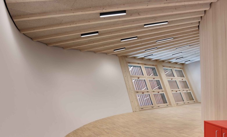 Heller, geschwungener Innenraum mit hellem Holzboden und einer Decke aus sichtbaren Holzbalken. Schlanke, schwarze Einbauleuchten sind regelmäßig zwischen den Balken angeordnet. Am Ende des Raums sorgt eine großflächige Holz-Glas-Konstruktion mit Lamellen für Tageslicht und eine warme, natürliche Atmosphäre.