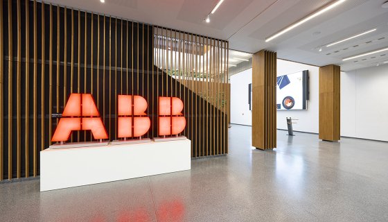 Im modernen Empfangsbereich eines Firmengebäudes steht ein leuchtend rotes ABB-Logo auf einem weißen Podest. Die Umgebung ist offen und hell gestaltet, mit Holzlamellen als Raumtrenner und digitalen Bildschirmen im Hintergrund.