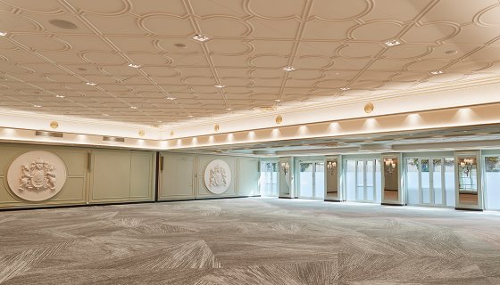 Prächtiger Ballsaal mit kunstvoll verzierter Decke, stilvollem Teppich und großflächigen Fenstern für natürliches Licht. Wandreliefs und klassische Leuchten unterstreichen das elegante Ambiente – ideal für festliche Anlässe, Galas oder Empfänge.