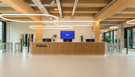 Modernes Empfangsdesign mit natürlichem Holz, offenen Sichtstrukturen und eleganter Lichtführung: Der Empfangsbereich von Universal Investment vereint Transparenz, Innovation und einladende Atmosphäre.