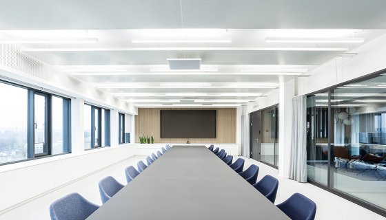 Moderner Konferenzraum mit großem, dunklem Besprechungstisch und blauen Stühlen. Helle, klare Architektur mit bodentiefen Fenstern sorgt für viel Tageslicht. Am Ende des Raumes befindet sich ein großer Bildschirm, flankiert von Holzpaneelen und Pflanzen. Rechtsseitig geben Glaswände den Blick in einen weiteren Raum frei.