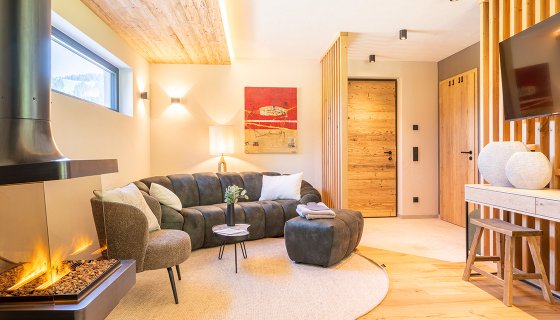 Ein gemütliches Wohnzimmer mit modernem Design: Ein Kamin mit warmem Feuer sorgt für eine behagliche Atmosphäre. Eine graue Eckcouch mit Kissen lädt zum Entspannen ein. Helles Holz an Wänden, Türen und Boden verleiht dem Raum eine natürliche Note. Dezente Beleuchtung und stilvolle Dekoration schaffen ein harmonisches, einladendes Ambiente.