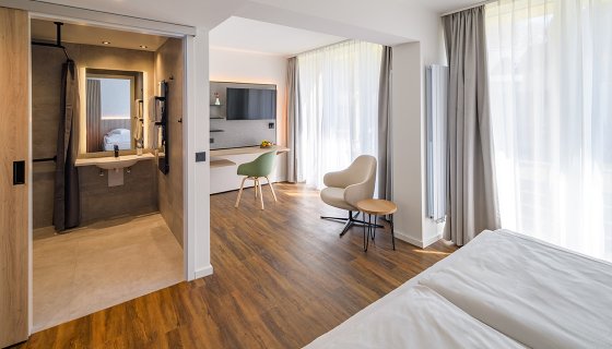 Helles, modern eingerichtetes Hotelzimmer mit warmem Holzboden, großem Bett und bodentiefen Fenstern mit Vorhängen. Ein offener Arbeitsbereich mit Schreibtisch, Stuhl und Wand-TV schließt an. Links ist ein offenes Bad mit Waschbecken, Spiegel und Dusche zu sehen. Die Einrichtung wirkt ruhig, hochwertig und einladend.