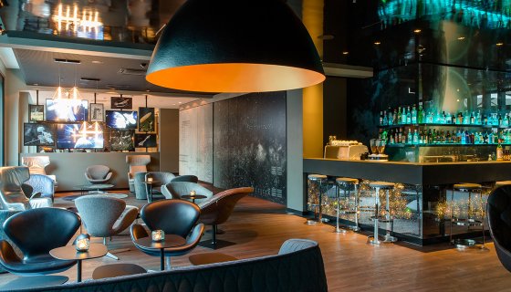 Moderne Bar- und Loungebereich mit eleganter Innenarchitektur. Gepolsterte Sessel und Sofas sind um niedrige Tische gruppiert, darüber hängen große, dunkle Design-Leuchten mit warmem Licht. Im Hintergrund befindet sich eine beleuchtete Bar mit Glasregalen und Spirituosen. Holzfußboden, dunkle Farben und stimmungsvolle Beleuchtung schaffen eine exklusive, gemütliche Atmosphäre.
