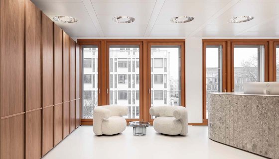 Modern gestalteter Empfangsbereich mit warmen Holzverkleidungen an der Wand, großen bodentiefen Fenstern und hellem Boden. Zwei weiche, abgerundete Lounge-Sessel stehen um einen kleinen Tisch. Rechts befindet sich eine halbkreisförmige Empfangstheke aus Stein. Das Tageslicht sorgt für eine ruhige, einladende Atmosphäre.