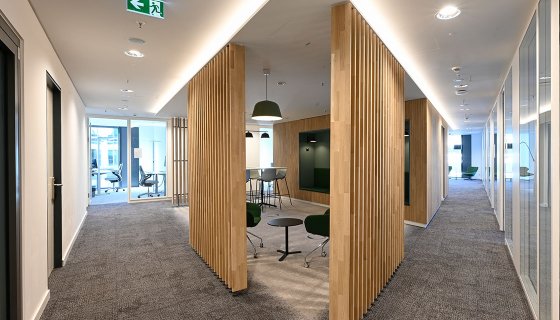 Moderner Büroflur mit grauem Teppichboden und hellen Wänden. In der Mitte stehen raumteilende Holzlamellen-Elemente, die kleine Besprechungsbereiche mit Sitzmöbeln und Tischen bilden. Rechts verläuft ein Gang mit verglasten Büros, links führen Türen zu weiteren Räumen. Dezente Deckenbeleuchtung sorgt für eine helle, freundliche Atmosphäre.