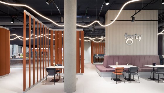 Modern gestalteter Innenraum eines Restaurants oder Cafés mit minimalistischer Einrichtung: Helle Böden, offene Decke mit geschwungenen Lichtleisten, orangefarbene vertikale Raumteiler und eine große Sitzbank in Rosé. Mehrere kleine Tische mit Stühlen sind verteilt. Eine Betonstütze steht zentral, im Hintergrund ist eine Bar sichtbar. An der Wand steht der Schriftzug „forty“.