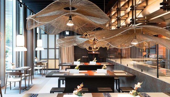 Modern gestalteter Restaurantbereich mit großzügigen Gemeinschaftstischen in einem hellen, industriell-eleganten Ambiente. Über den Tischen schweben organisch geformte Deckenelemente aus filigranem, netzartigem Material, die an Wellen erinnern. Große Fensterflächen, warme Pendelleuchten und dezente florale Dekorationen schaffen eine einladende Atmosphäre. Die Kombination aus Metall, Holz und Naturtönen vermittelt Modernität und hochwertige Gestaltung.
