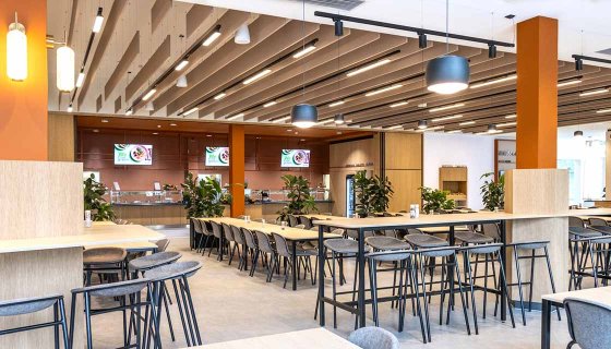 Moderne, offen gestaltete Kantine mit heller und freundlicher Atmosphäre. Großzügige Sitzbereiche, hohe Tische und eine zeitgemäße Einrichtung schaffen ideale Bedingungen für entspannte Pausen und den Austausch unter Kolleginnen und Kollegen. Warme Holzelemente, dezente Farben und zahlreiche Pflanzen sorgen für ein einladendes Ambiente und ein angenehmes Wohlfühlklima.