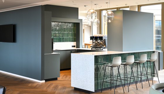 Moderne Bürolounge mit offenem Küchenbereich und stilvoller Barinsel. Hochwertige Materialien wie Holz, Naturstein und grüne Fliesen schaffen eine einladende Atmosphäre. Großzügige Fenster sorgen für viel Tageslicht, während elegante Pendelleuchten und zeitloses Design den Raum zu einem idealen Treffpunkt für Pausen und informellen Austausch machen.