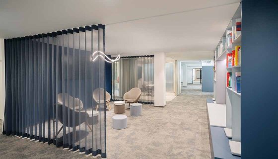 Moderner, offen gestalteter Bürobereich mit hellen Teppichböden und einer transparenten Lamellenabtrennung, die Besprechungszonen dezent voneinander trennt. Bequeme Loungesessel und Hocker schaffen eine einladende Atmosphäre, während offene Regale und großzügige Laufwege Funktionalität und Übersichtlichkeit verbinden. Das helle Design fördert Kommunikation, Konzentration und flexibles Arbeiten.