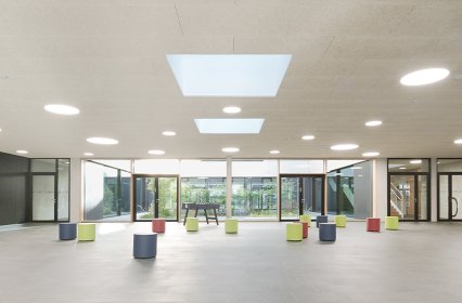 Großer, lichtdurchfluteter Innenraum mit modernen Designelementen. Die Decke verfügt über mehrere runde Leuchten und zwei große quadratische Oberlichter, die natürliches Licht hereinlassen. Vor einer Glasfassade mit Blick auf einen begrünten Außenbereich stehen farbenfrohe, zylinderförmige Sitzhocker in verschiedenen Farben wie Blau, Rot und Grün. Der Raum wirkt offen und minimalistisch, ideal für Begegnungen, Veranstaltungen oder gemeinschaftliche Aktivitäten.