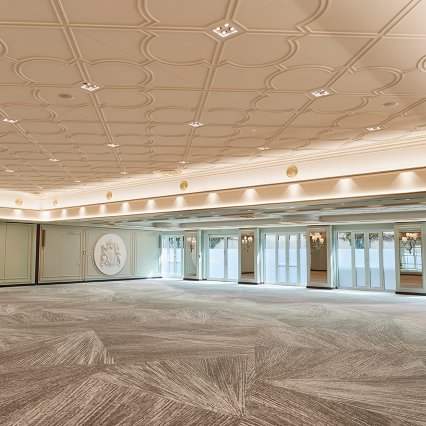 Großzügiger Ballsaal mit aufwendig gestalteter Decke, elegantem Teppichboden und großen Fensterfronten, die für viel Tageslicht sorgen. Wandreliefs mit Wappen sowie klassische Wandlampen verleihen dem Raum eine festliche und zugleich stilvolle Atmosphäre – ideal für exklusive Veranstaltungen, Bankette oder Galas.