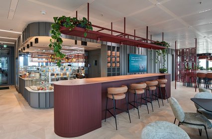 Modernes Café mit elegantem Design: Eine lange Theke mit Barhockern aus Holz und Leder, umgeben von warmen Rottönen und grünen Pflanzen. Im Hintergrund befindet sich eine Glasvitrine mit Gebäck und Snacks sowie ein gemütlicher Sitzbereich mit modernen Möbeln. Das Ambiente wirkt offen, einladend und stilvoll – ideal für entspannte Pausen oder informelle Gespräche.