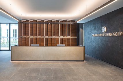 Modern gestalteter Empfangsbereich mit einer großen, hellen Empfangstheke im Vordergrund. Dahinter eine hohe Holzstruktur mit vertikalen Elementen und integrierter Beleuchtung. Rechts an der dunklen Wand befindet sich eine Uhr sowie ein Unternehmenslogo. Durch große Fenster fällt Tageslicht in den offenen, elegant eingerichteten Raum.