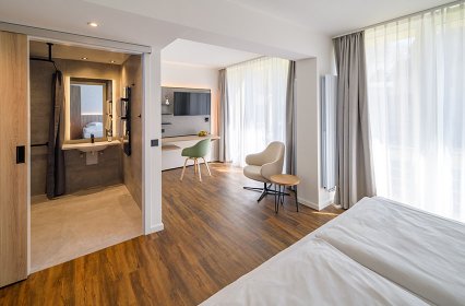 Helles, modern eingerichtetes Hotelzimmer mit warmem Holzboden, großem Bett und bodentiefen Fenstern mit Vorhängen. Ein offener Arbeitsbereich mit Schreibtisch, Stuhl und Wand-TV schließt an. Links ist ein offenes Bad mit Waschbecken, Spiegel und Dusche zu sehen. Die Einrichtung wirkt ruhig, hochwertig und einladend.