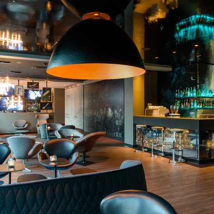Moderne Bar- und Loungebereich mit eleganter Innenarchitektur. Gepolsterte Sessel und Sofas sind um niedrige Tische gruppiert, darüber hängen große, dunkle Design-Leuchten mit warmem Licht. Im Hintergrund befindet sich eine beleuchtete Bar mit Glasregalen und Spirituosen. Holzfußboden, dunkle Farben und stimmungsvolle Beleuchtung schaffen eine exklusive, gemütliche Atmosphäre.
