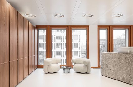 Modern gestalteter Empfangsbereich mit warmen Holzverkleidungen an der Wand, großen bodentiefen Fenstern und hellem Boden. Zwei weiche, abgerundete Lounge-Sessel stehen um einen kleinen Tisch. Rechts befindet sich eine halbkreisförmige Empfangstheke aus Stein. Das Tageslicht sorgt für eine ruhige, einladende Atmosphäre.