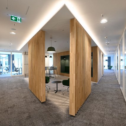 Moderner Büroflur mit grauem Teppichboden und hellen Wänden. In der Mitte stehen raumteilende Holzlamellen-Elemente, die kleine Besprechungsbereiche mit Sitzmöbeln und Tischen bilden. Rechts verläuft ein Gang mit verglasten Büros, links führen Türen zu weiteren Räumen. Dezente Deckenbeleuchtung sorgt für eine helle, freundliche Atmosphäre.