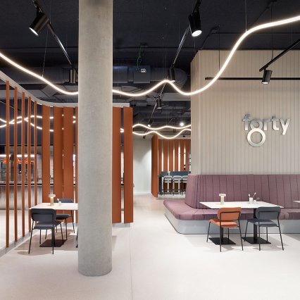 Modern gestalteter Innenraum eines Restaurants oder Cafés mit minimalistischer Einrichtung: Helle Böden, offene Decke mit geschwungenen Lichtleisten, orangefarbene vertikale Raumteiler und eine große Sitzbank in Rosé. Mehrere kleine Tische mit Stühlen sind verteilt. Eine Betonstütze steht zentral, im Hintergrund ist eine Bar sichtbar. An der Wand steht der Schriftzug „forty“.