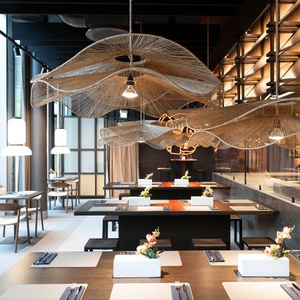 Modern gestalteter Restaurantbereich mit großzügigen Gemeinschaftstischen in einem hellen, industriell-eleganten Ambiente. Über den Tischen schweben organisch geformte Deckenelemente aus filigranem, netzartigem Material, die an Wellen erinnern. Große Fensterflächen, warme Pendelleuchten und dezente florale Dekorationen schaffen eine einladende Atmosphäre. Die Kombination aus Metall, Holz und Naturtönen vermittelt Modernität und hochwertige Gestaltung.